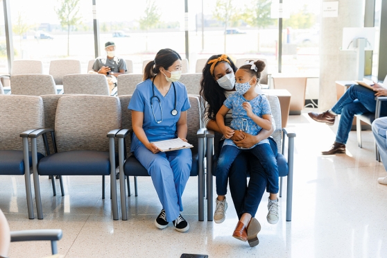 Un niño se sienta en el regazo de un adulto junto a un médico. Todos llevan máscaras.
